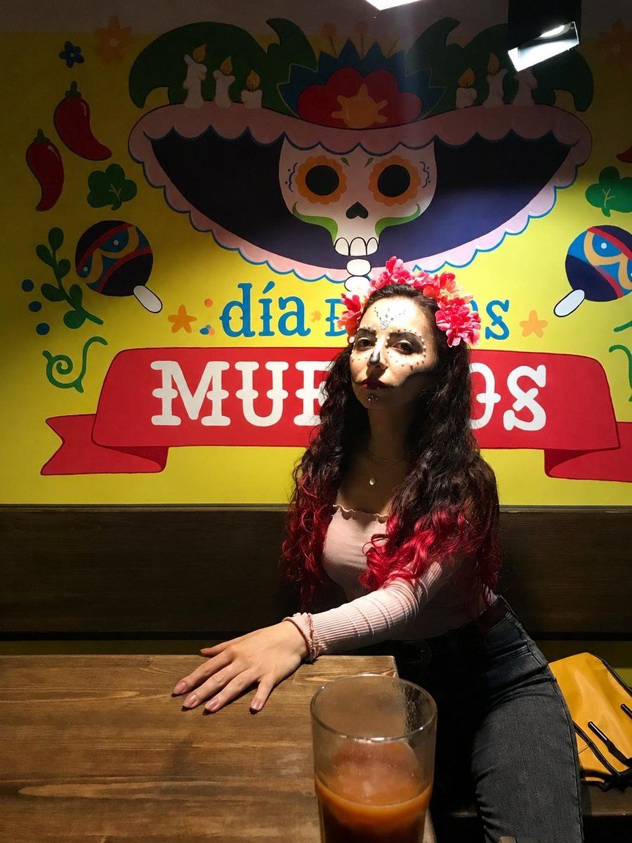 Naira in Día de Muertos make-up with a flower crown, in front of a Día de los Muertos mural