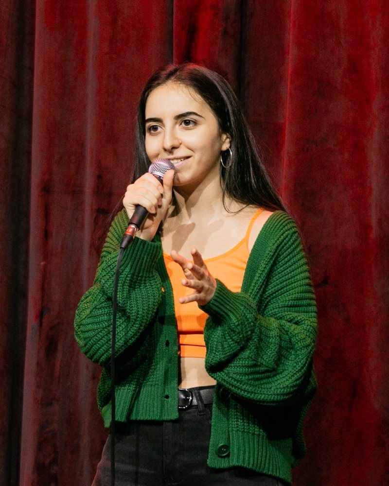 Naira performing stand-up with a microphone in front of a red curtain